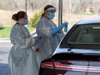 Two nurses dressed in PPE at a Saint Luke's drive-thru testing site, talking with a patient through their car window holding sampling collection kit