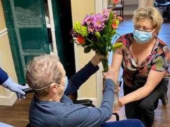 Anita, sitting in a wheel chair, puts flowers in a vase held by a member of her care team