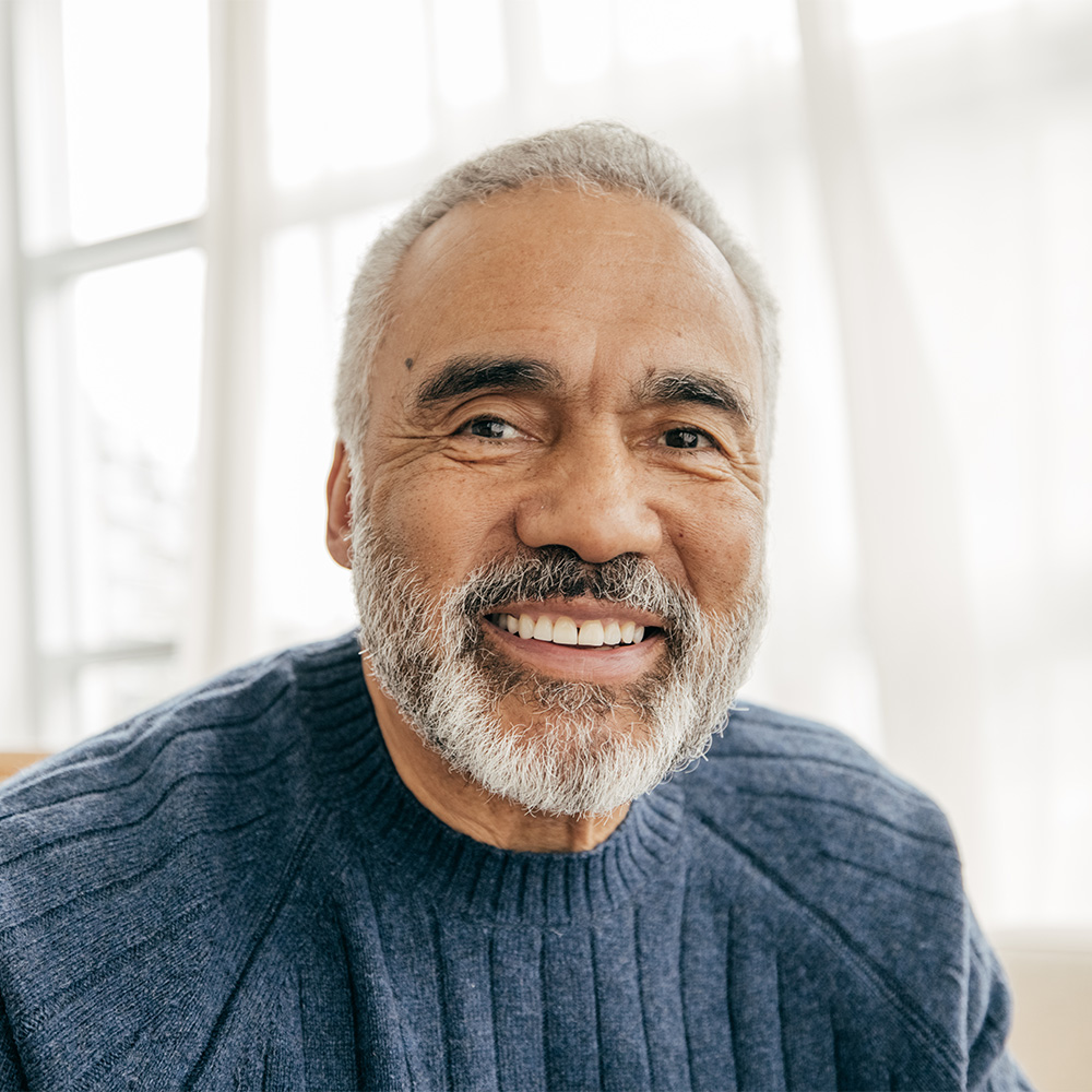 A smiling man with a grey beard in a blue sweatshirt in a bright room with windows.