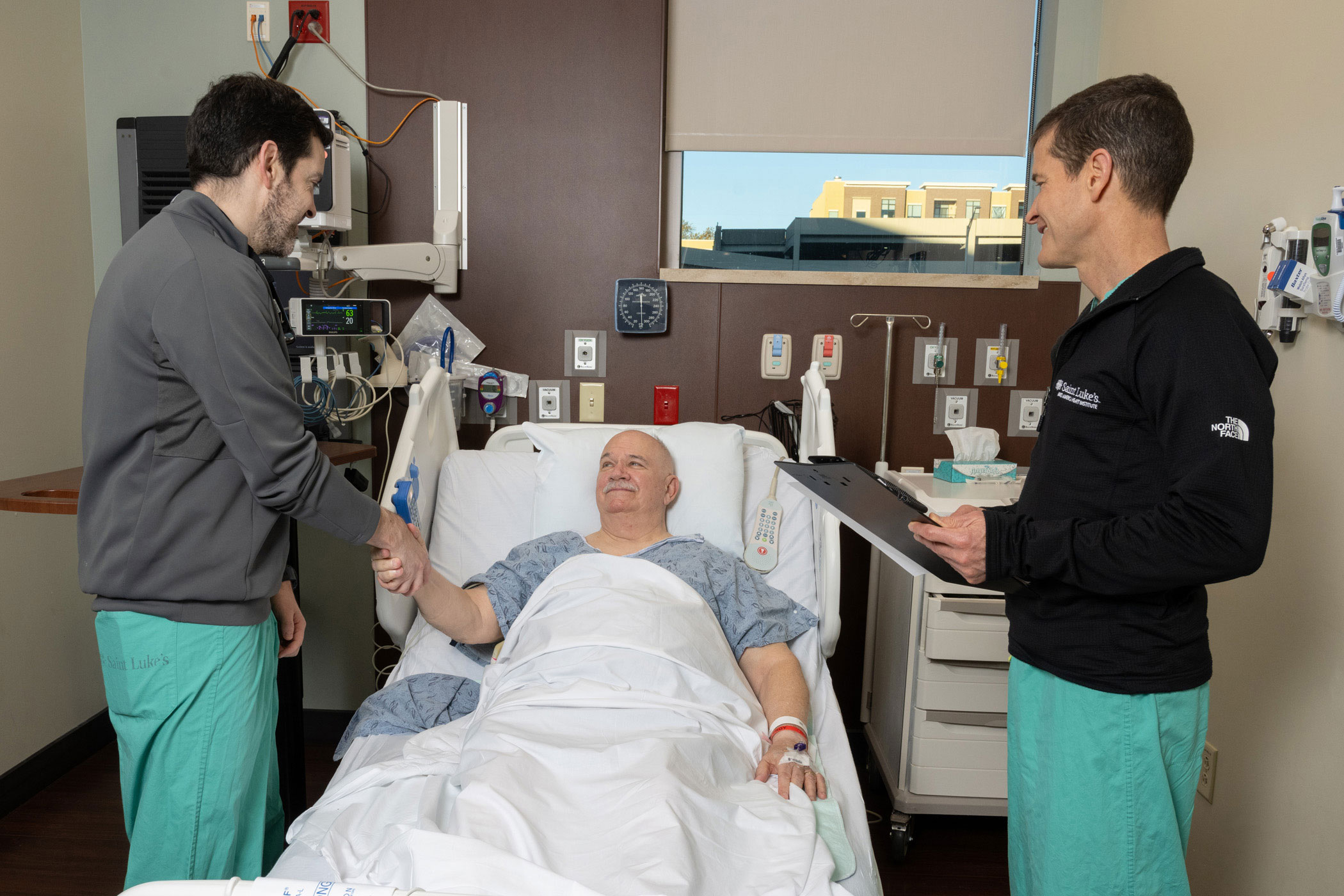 Two Saint Luke's cardiologists are standing around a smiling patient in a hospital bed.