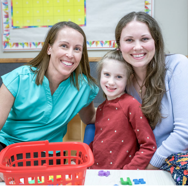 A speech therapist is smiling into the camera with a child and her mother.