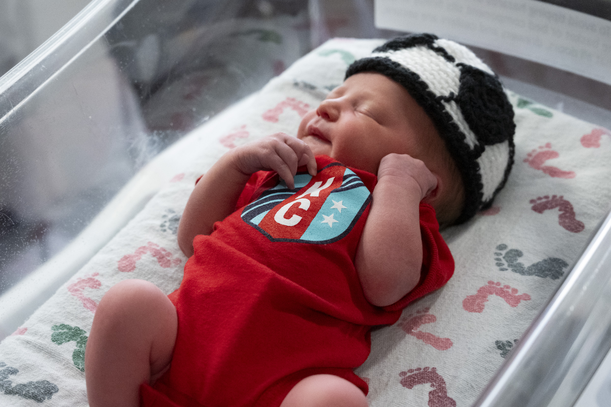 A baby wearing a red KC Current onesie, laying in a hospital bed