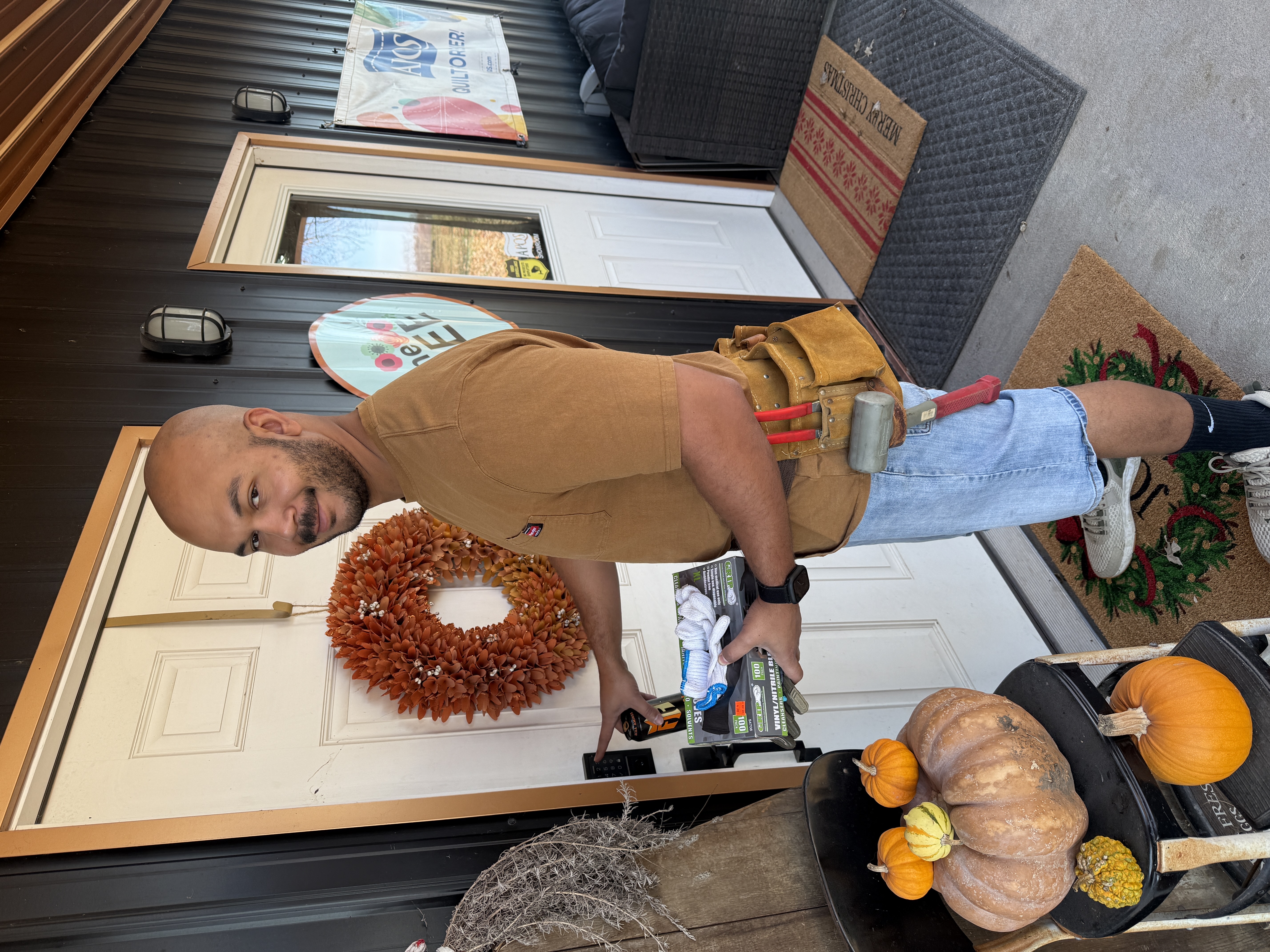 Justin in a light brown shirt and denim shorts, standing near a white door with an orange wreath and pumpkins on the floor