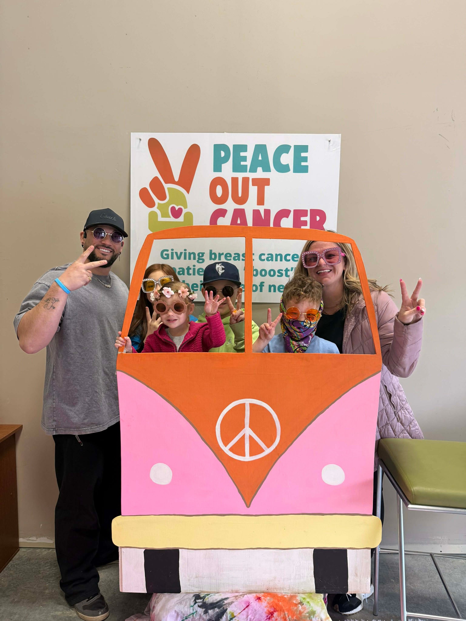 Karissa (right) and her husband and four children all wearing costume sunglasses, posing behind a painted cardboard bus structure with a peace sign and a sign behind them saying "Peace out Cancer!"