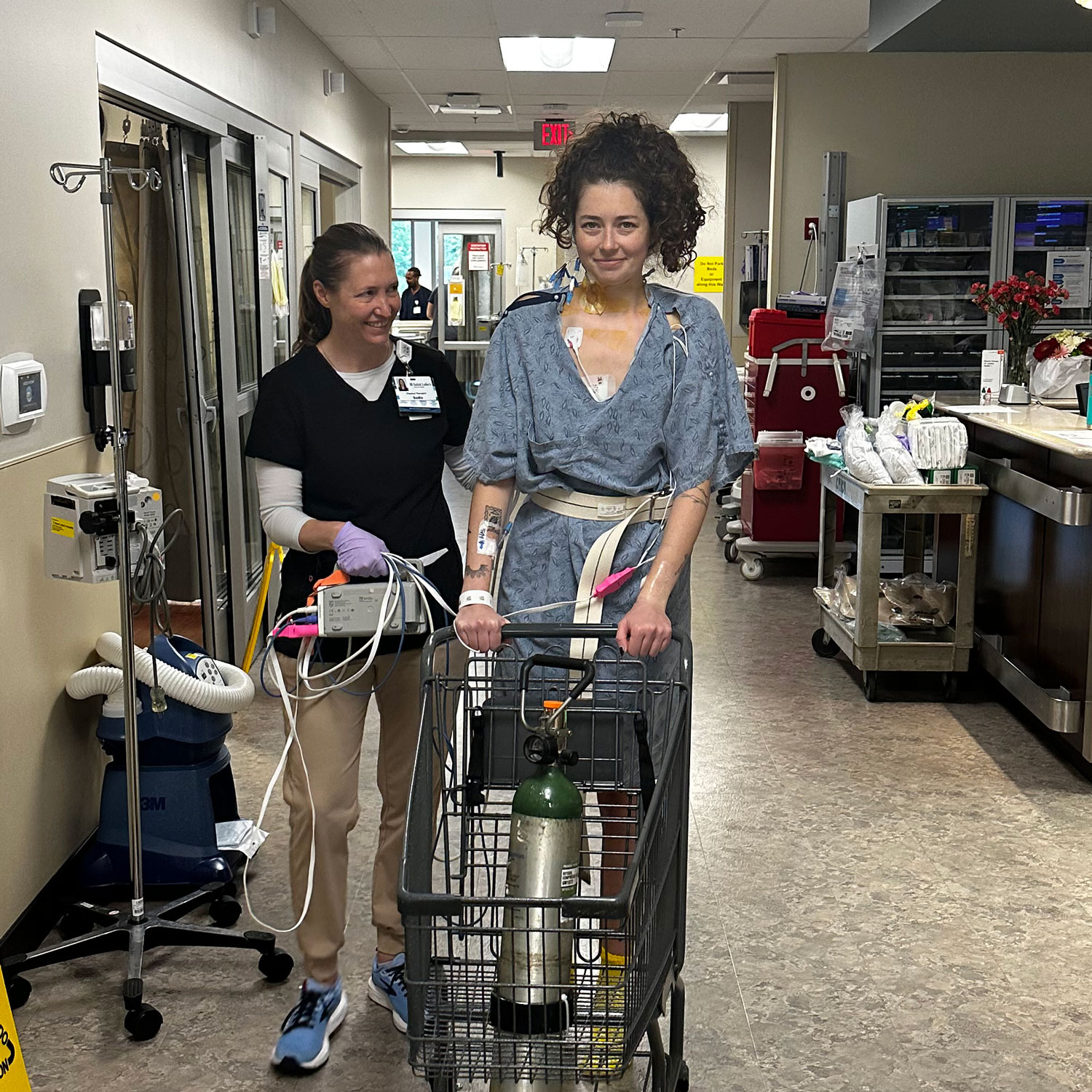Katie, a hospital patient in a gown, is being helped walking with a health care provider.