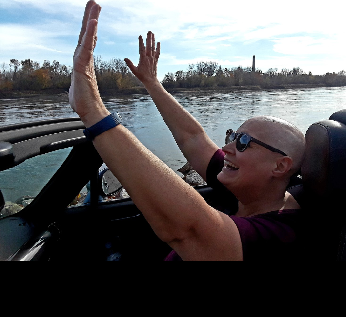 Lori is smiling, lifting her hands to the air in front of a beautiful river scene.