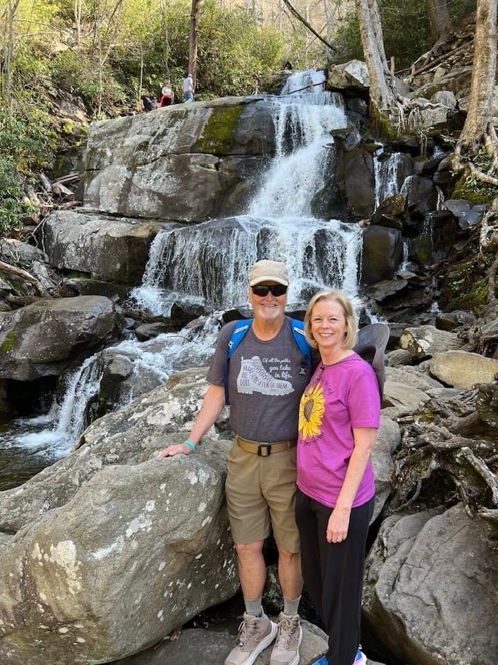 Joe (left) in a grey shirt with white text and graphic, standing near his wife (blonde hair and pink shirt), both in front of a waterfall