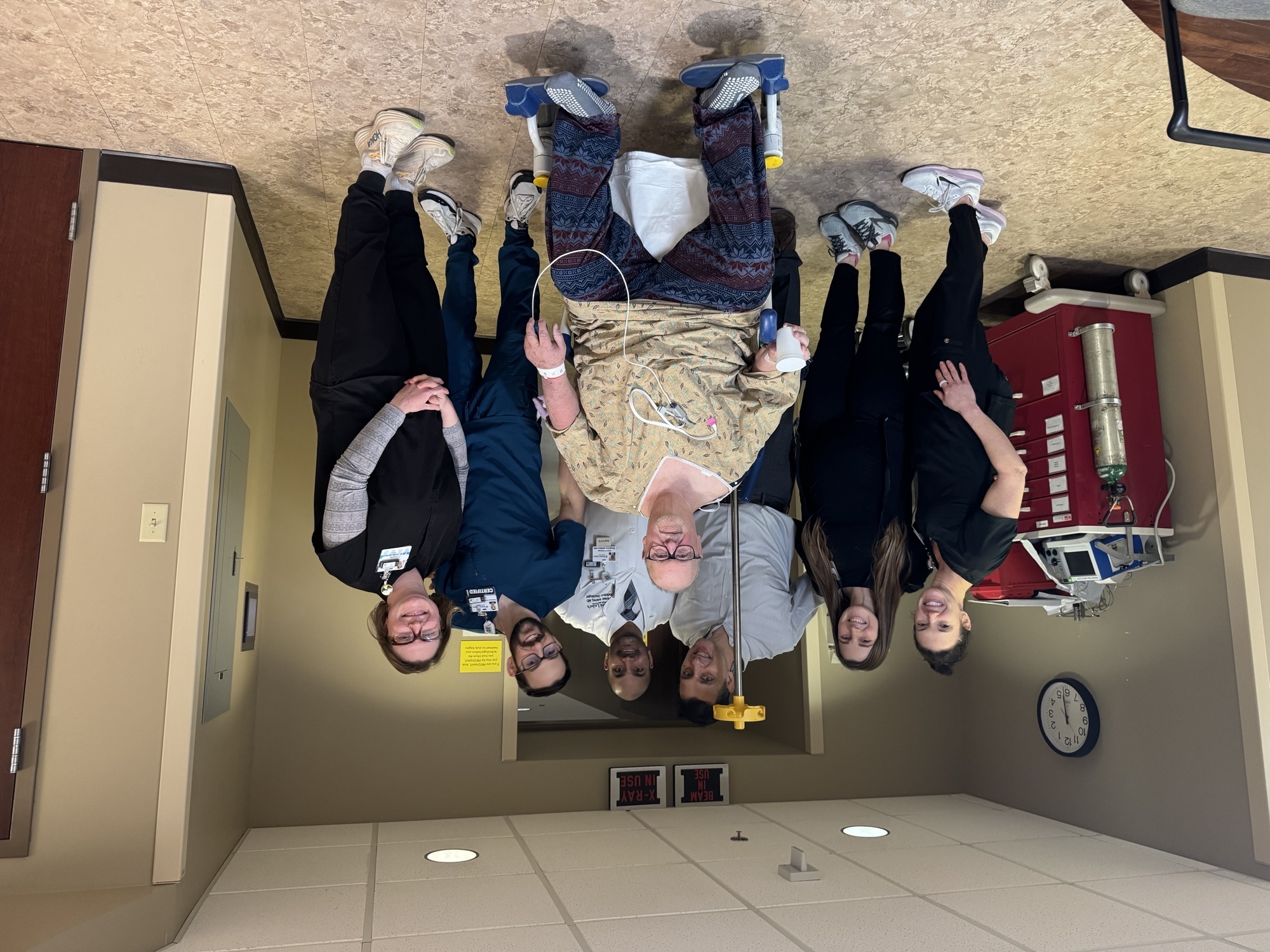 Noah Shoots (middle) in hospital scrubs, sitting on a wheelchair, in front of a five person care team, all wearing scrubs, standing behind in a hospital hallway.